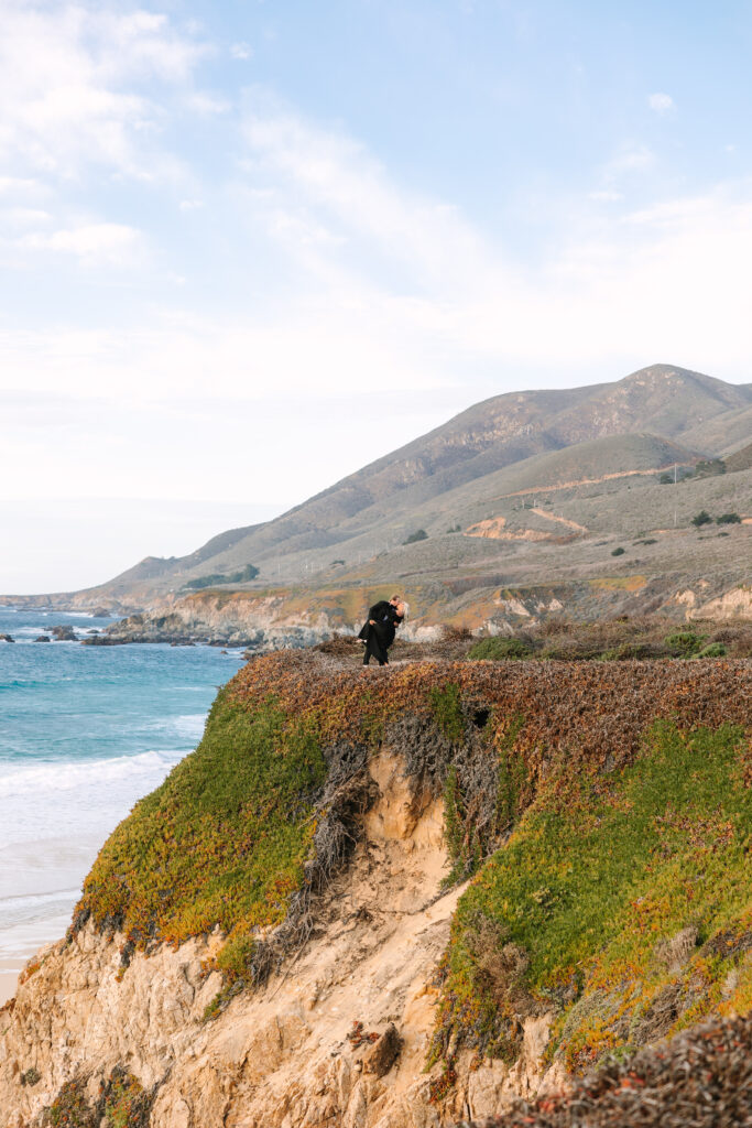 A couple sharing a tender kiss on a cliff in Big Sur, with the breathtaking coastline and ocean in the background. The warm glow of the setting sun casts a romantic light over the scene, highlighting the couple's intimate moment amidst the dramatic natural beauty of the rugged cliffs and vast ocean