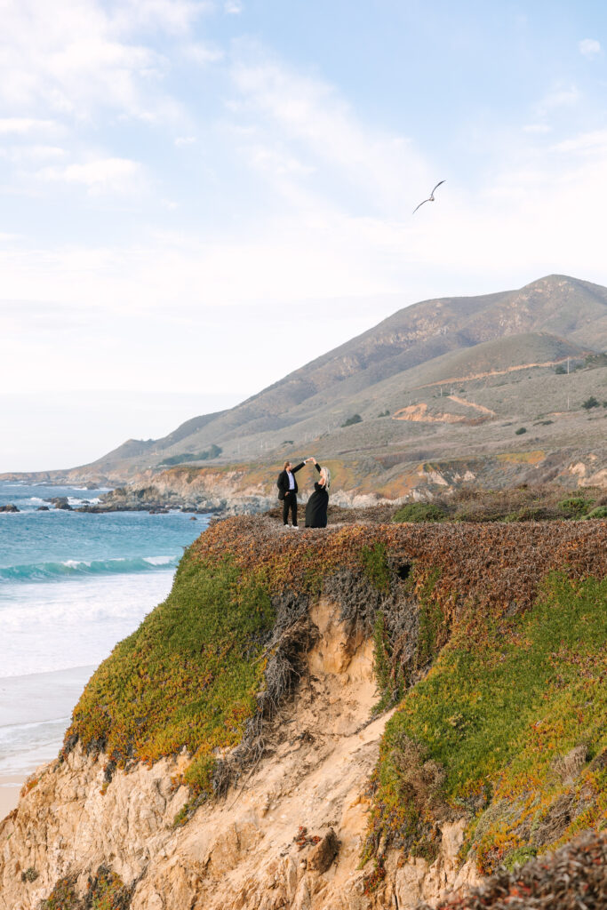 A couple standing on a cliff overlooking the dramatic coastline of Big Sur during a surprise proposal. The person proposing is on one knee, holding a ring, while their partner looks surprised and emotional. The sun sets in the background, casting a warm golden light over the breathtaking scenery of cliffs, ocean, and rugged mountains.