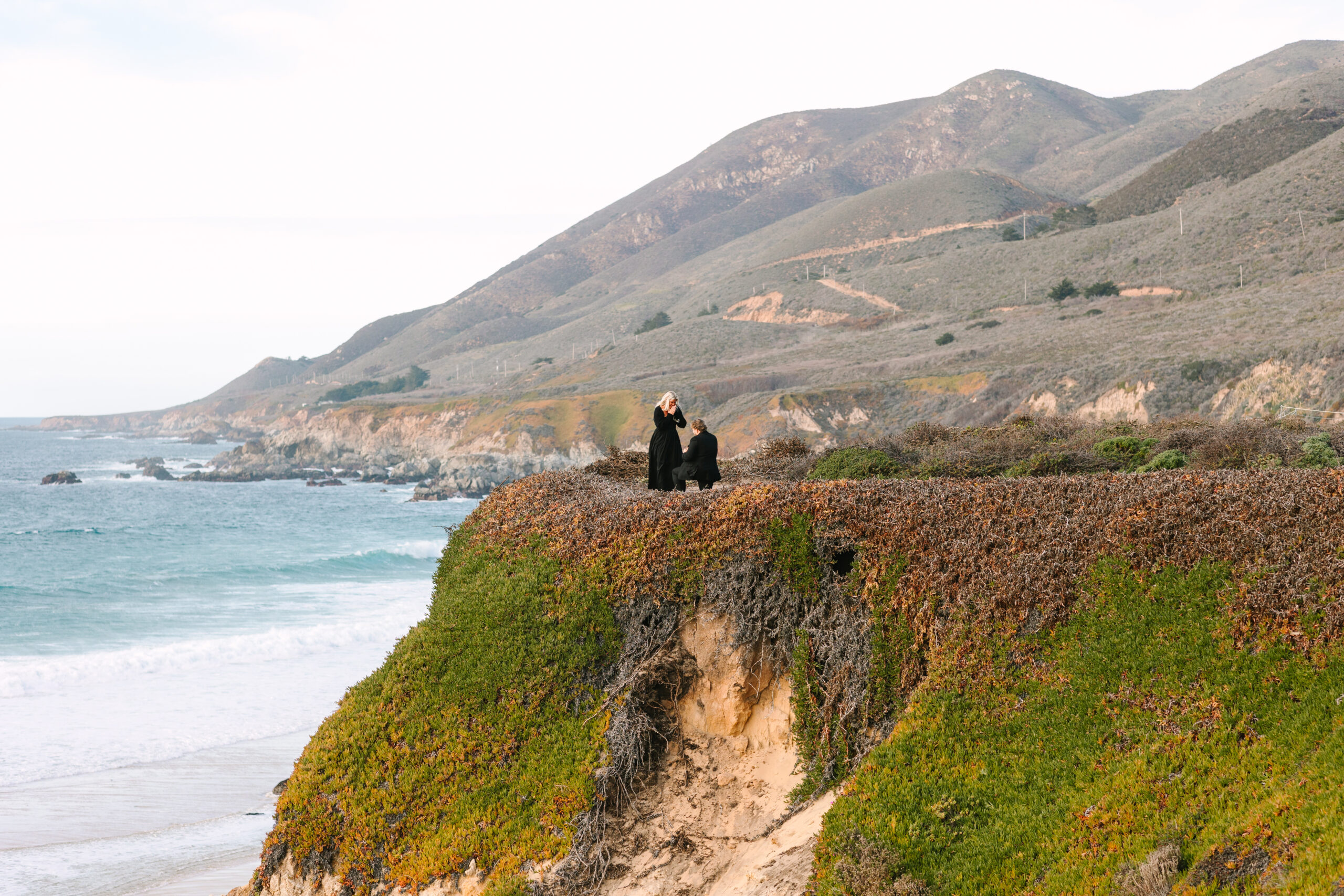 A couple standing on a cliff overlooking the dramatic coastline of Big Sur during a surprise proposal. The person proposing is on one knee, holding a ring, while their partner looks surprised and emotional. The sun sets in the background, casting a warm golden light over the breathtaking scenery of cliffs, ocean, and rugged mountains