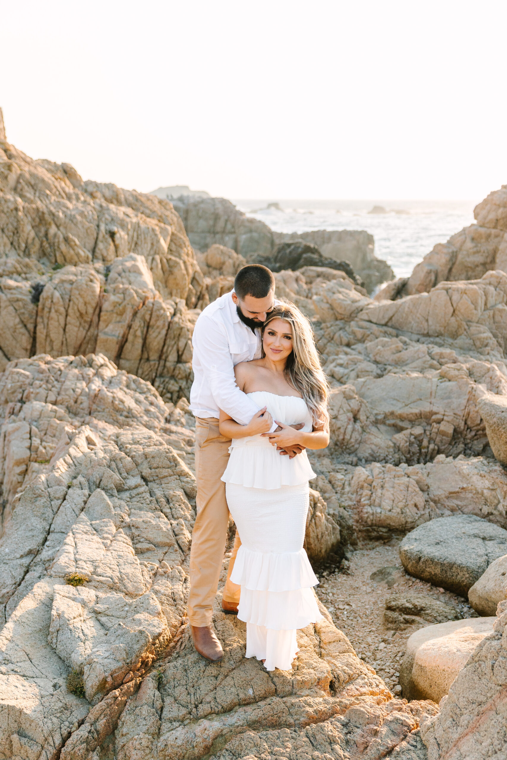 Couple posing during a romantic Big Sur engagement session, surrounded by scenic cliffs and the beautiful California coastline. The woman is wearing a flowing white dress, and the man is dressed in a neutral outfit, complementing the natural beauty of the landscape