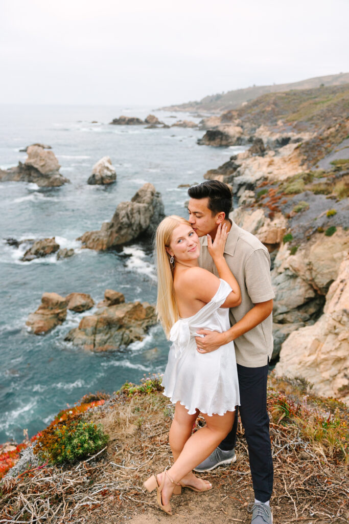 man kissing his fiance on her temple with stunning Big Sur views